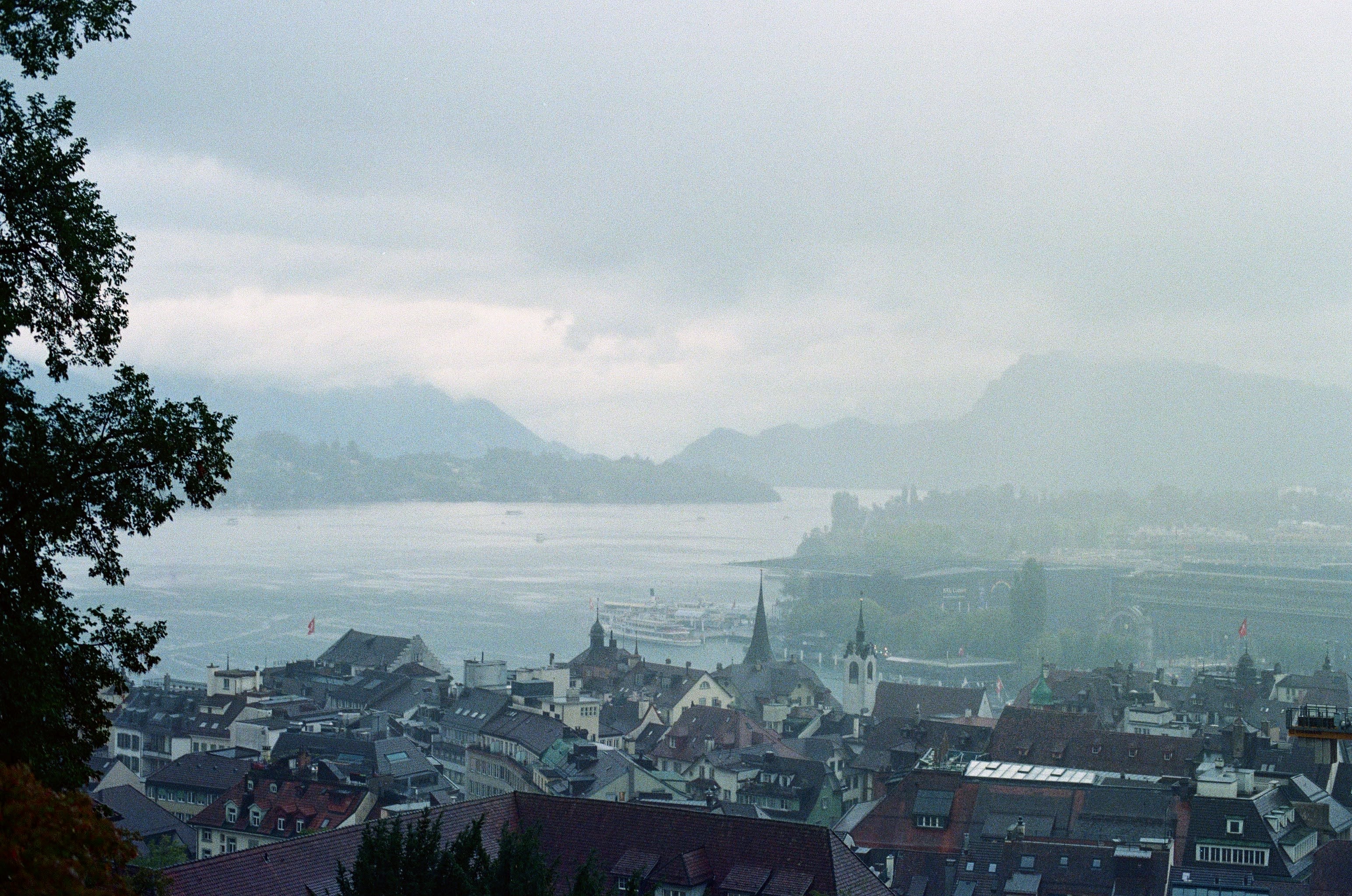 Rainy clouds cover half the frame which overlooks a city and a lake with mountains in the background.