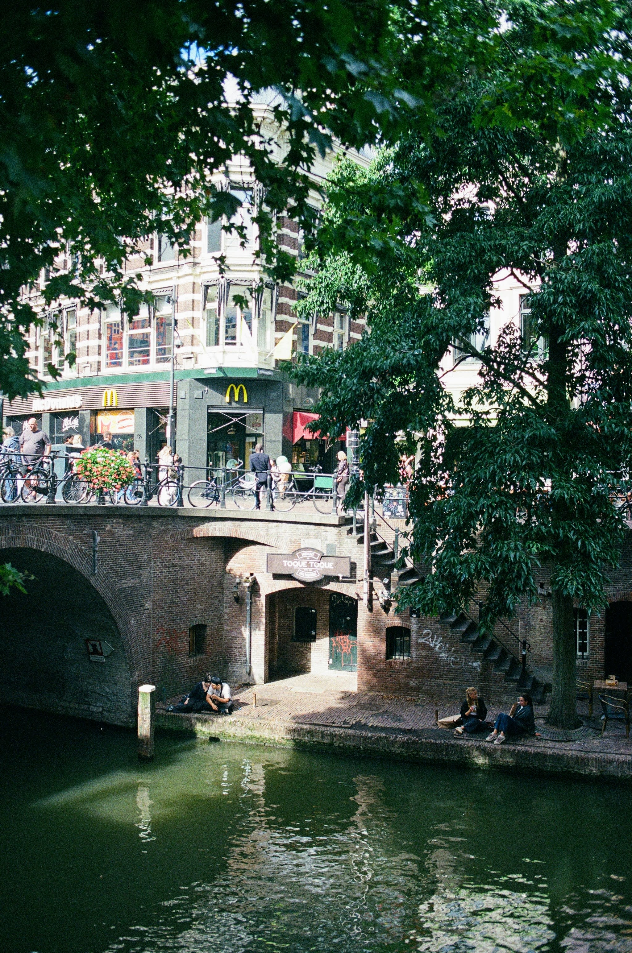 People sit in the shade by a canal underneath a busy road. A McDonald's restaurant is in the background, while the canal bank has a narrow sliver of sunshine that is being cast on it. 