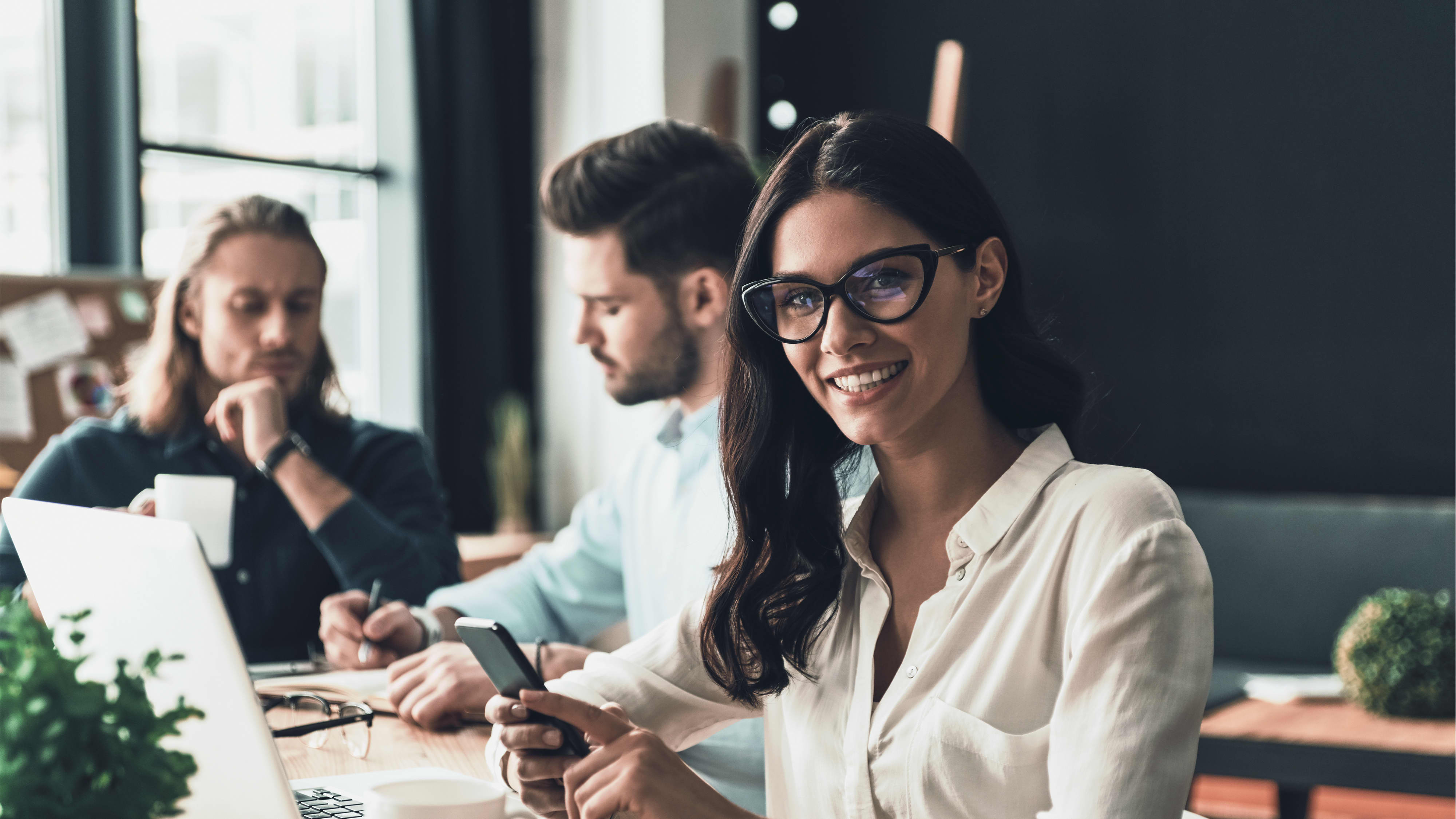 Frau arbeitet mit Smartphone und Laptop in modernem Büro