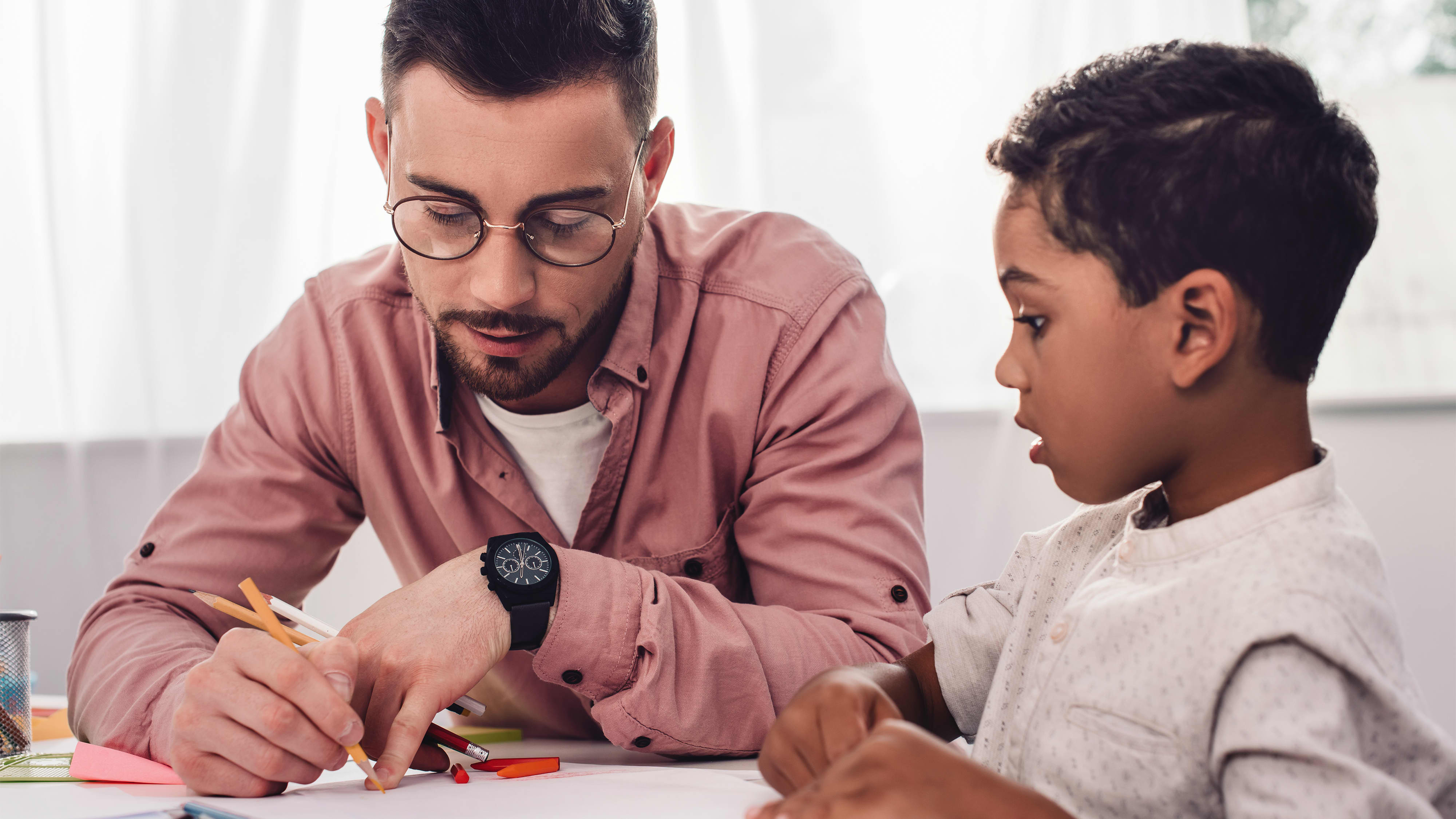 Karrieremöglichkeit “Sozialarbeiter:in im Bereich Schule”: Junger Sozialarbeiter mit Brille sitzt mit einem Grundschulkind am Tisch und unterstützt ihn bei Aufgaben