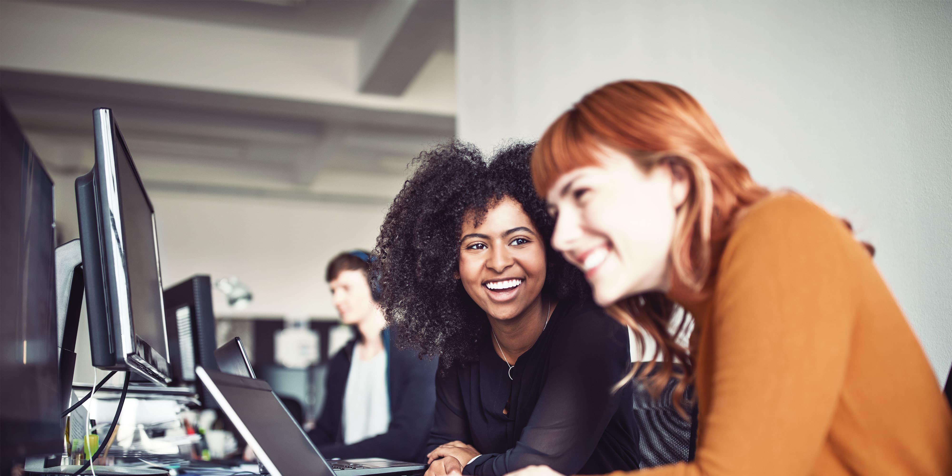 Karrieremöglichkeit “Technische:r Projektleiter:in”: Technische Projektleiterin arbeitet im Büro zusammen mit einer Kollegin aus dem Content-Team. Sie sitzen vor einem Laptop und lachen