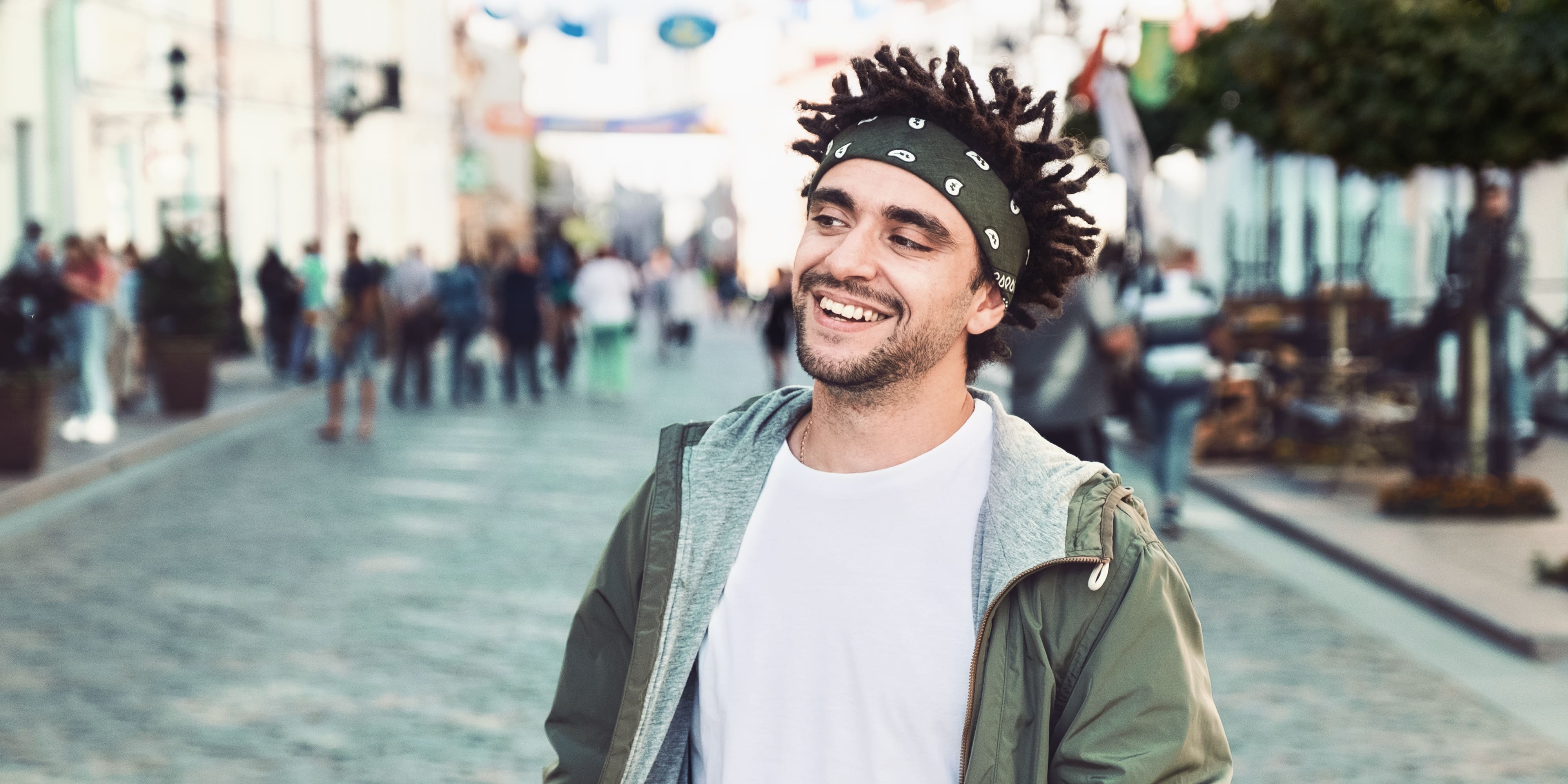 Junge stattliche Bart Mann gekleidet stilvolle lässige Kleidung, weißes T-Shirt, grüne Jacke, Bandana posiert im Freien. Lächelnde Hipster mit Dreadlocks Frisur, Nahaufnahme Porträt, Straße Foto, urbanen Stil