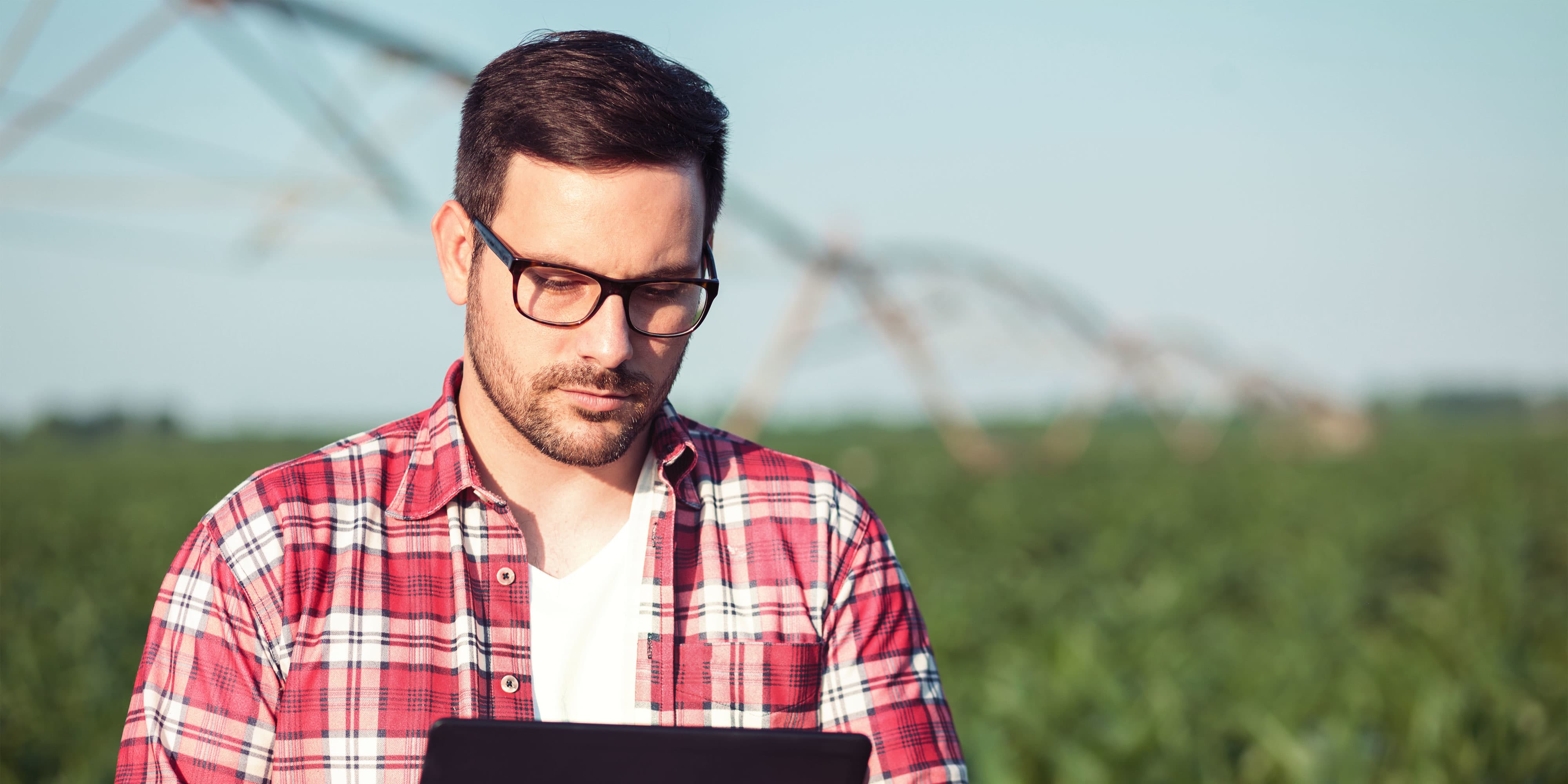 Karrieremöglichkeit “Produktmanager:in im Agrarhandel”: Produktmanager mit Brille und kariertem Hemd steht vor einem Feld und schaut auf sein Tablet