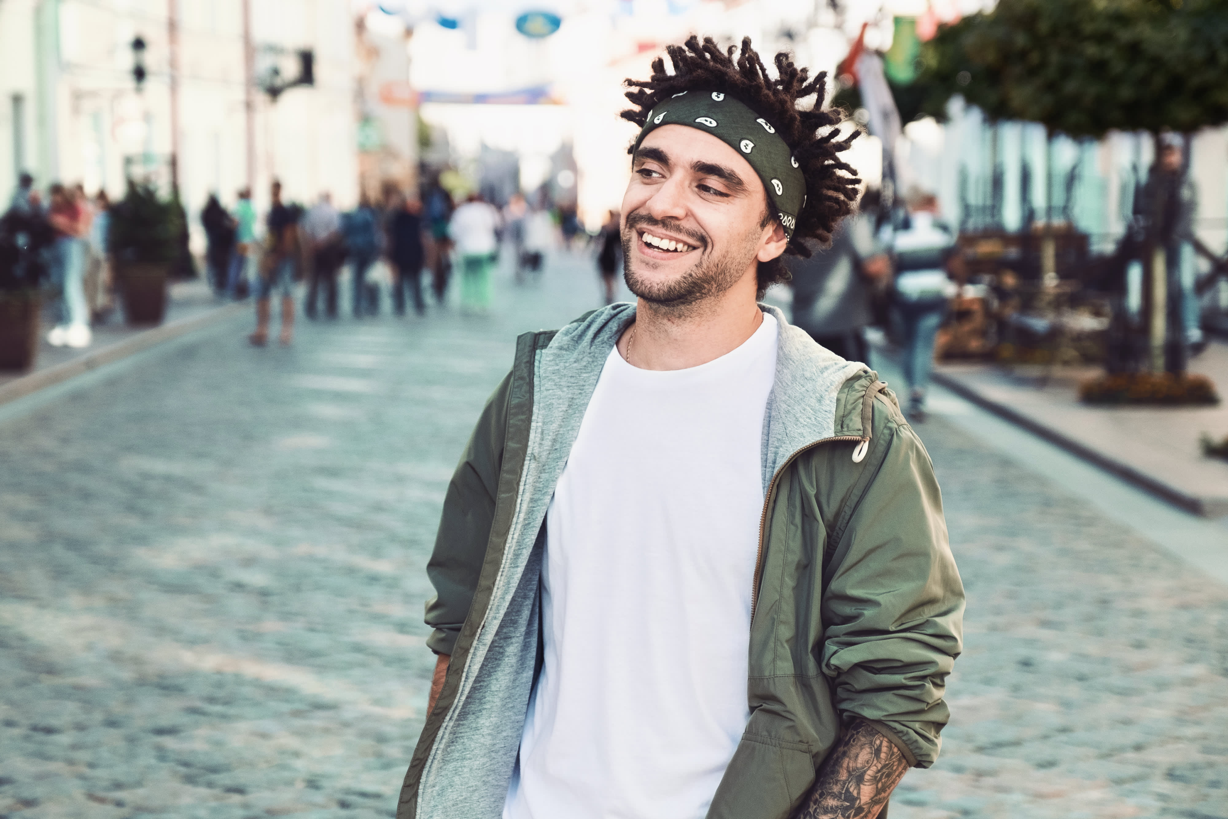 Junge stattliche Bart Mann gekleidet stilvolle lässige Kleidung, weißes T-Shirt, grüne Jacke, Bandana posiert im Freien. Lächelnde Hipster mit Dreadlocks Frisur, Nahaufnahme Porträt, Straße Foto, urbanen Stil