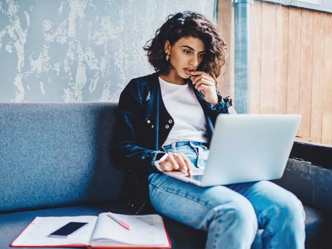 Duale Studentin sitzt auf dem Sofa mit ihrem Laptop und möchte ihr altes duales Studium abbrechen und an der IU Duales Studium neu starten.