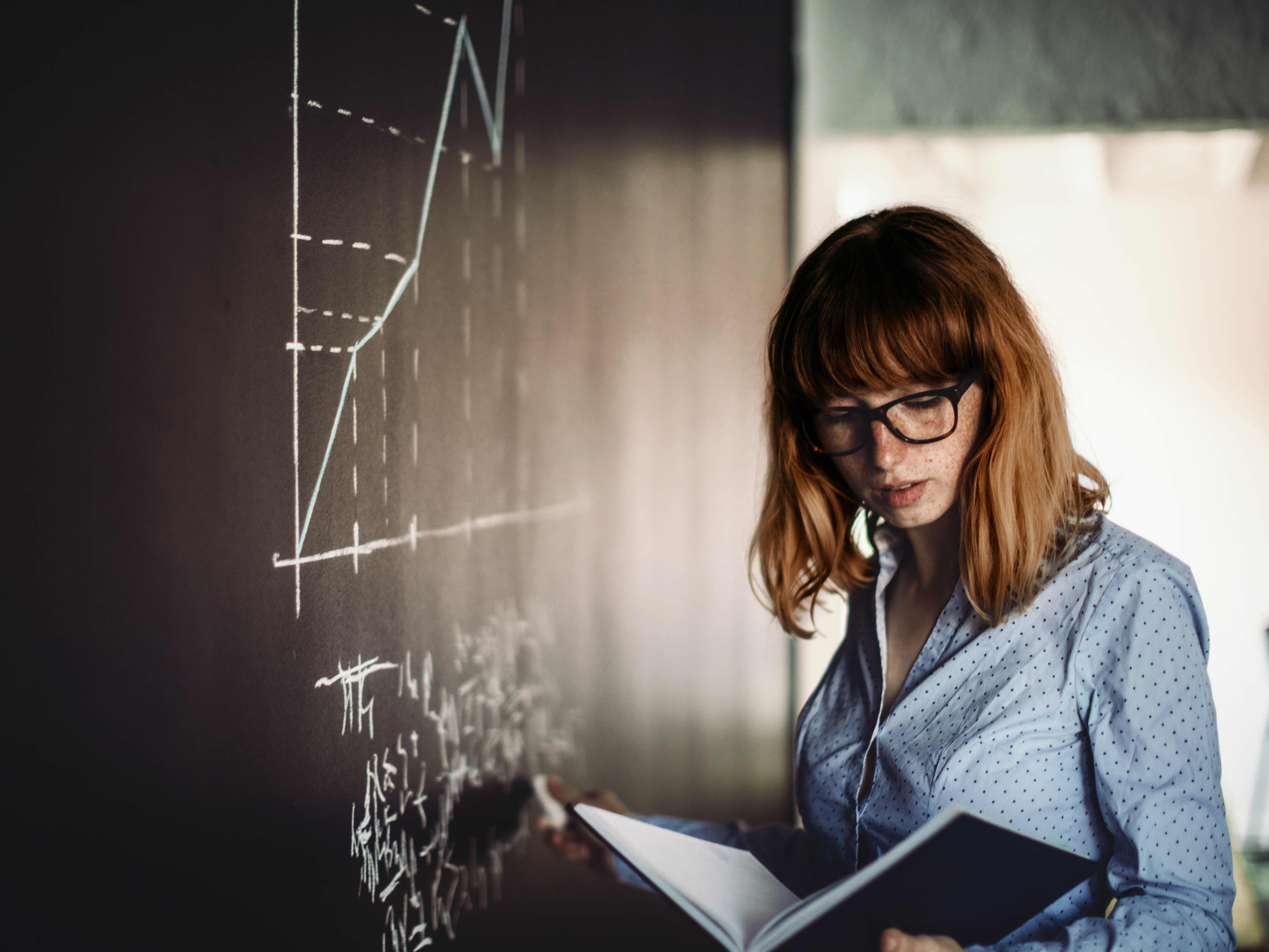 Titelbild der Infoseite "Duales Studium Wirtschaft": Duale Wirtschaftsstudentin steht an der Tafel mit einem Buch und Kreide in der Hand. Auf der Tafel sind Formeln und ein Graph zu sehen.
