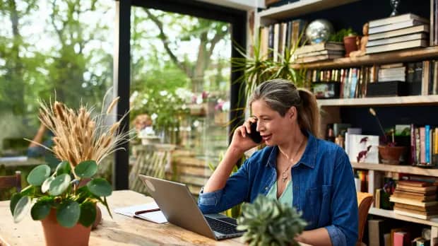 Eine Frau schaut auf ihren Laptop und telefoniert, im Hintergrund sieht man ihre Wohnung