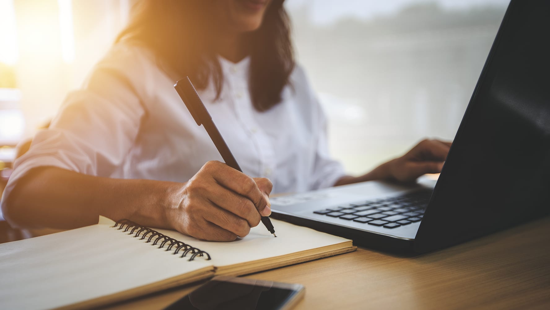 A woman in a white blouse sits at a desk, taking notes in a notebook while using a laptop. Warm sunlight streams through a window, casting a soft glow on her workspace.