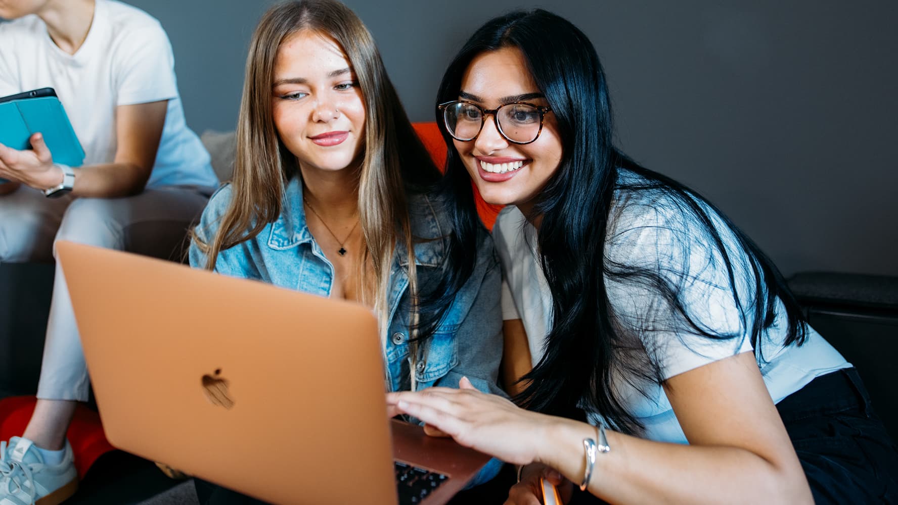 Two female students are smiling and looking together at a laptop screen on the IU Cologne campus.