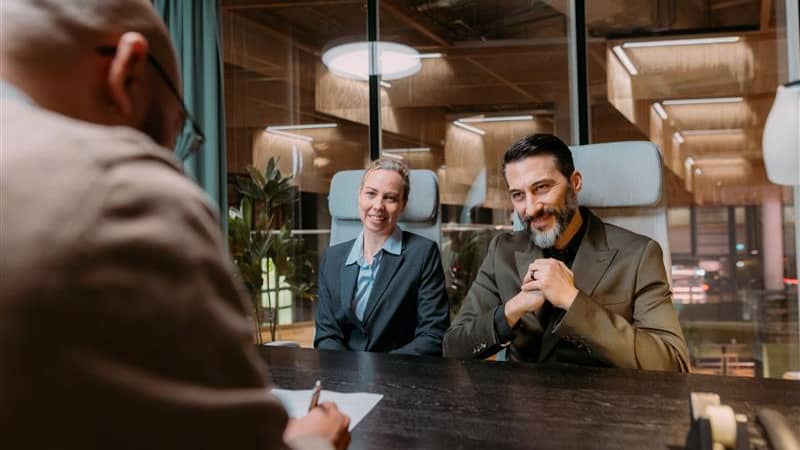 Two employers sit and smile to a new employee who sings a job contract in an office.