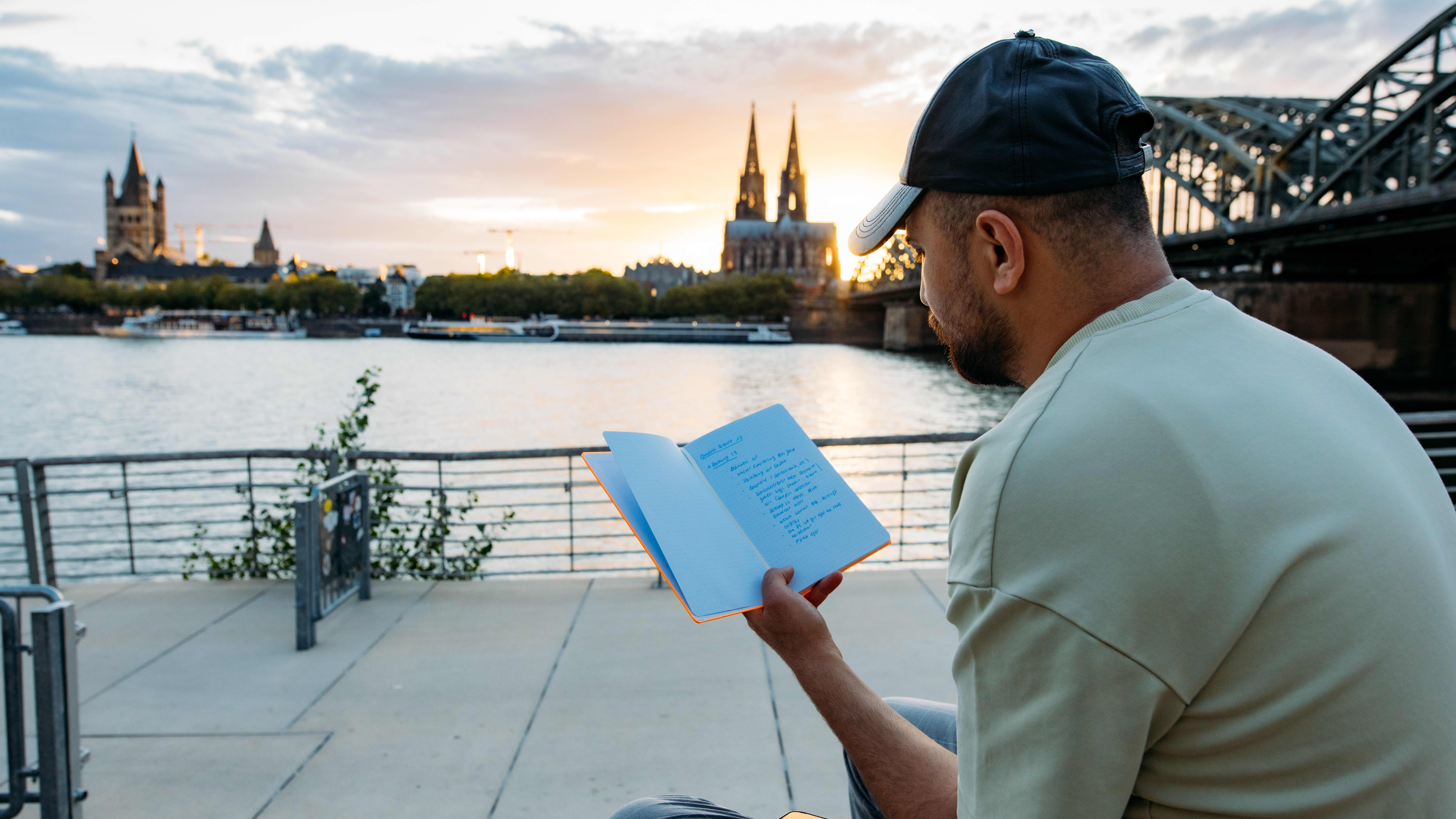 Man sitting and reading a book at sunset in Cologne, with the Cologne Cathedral in the background.