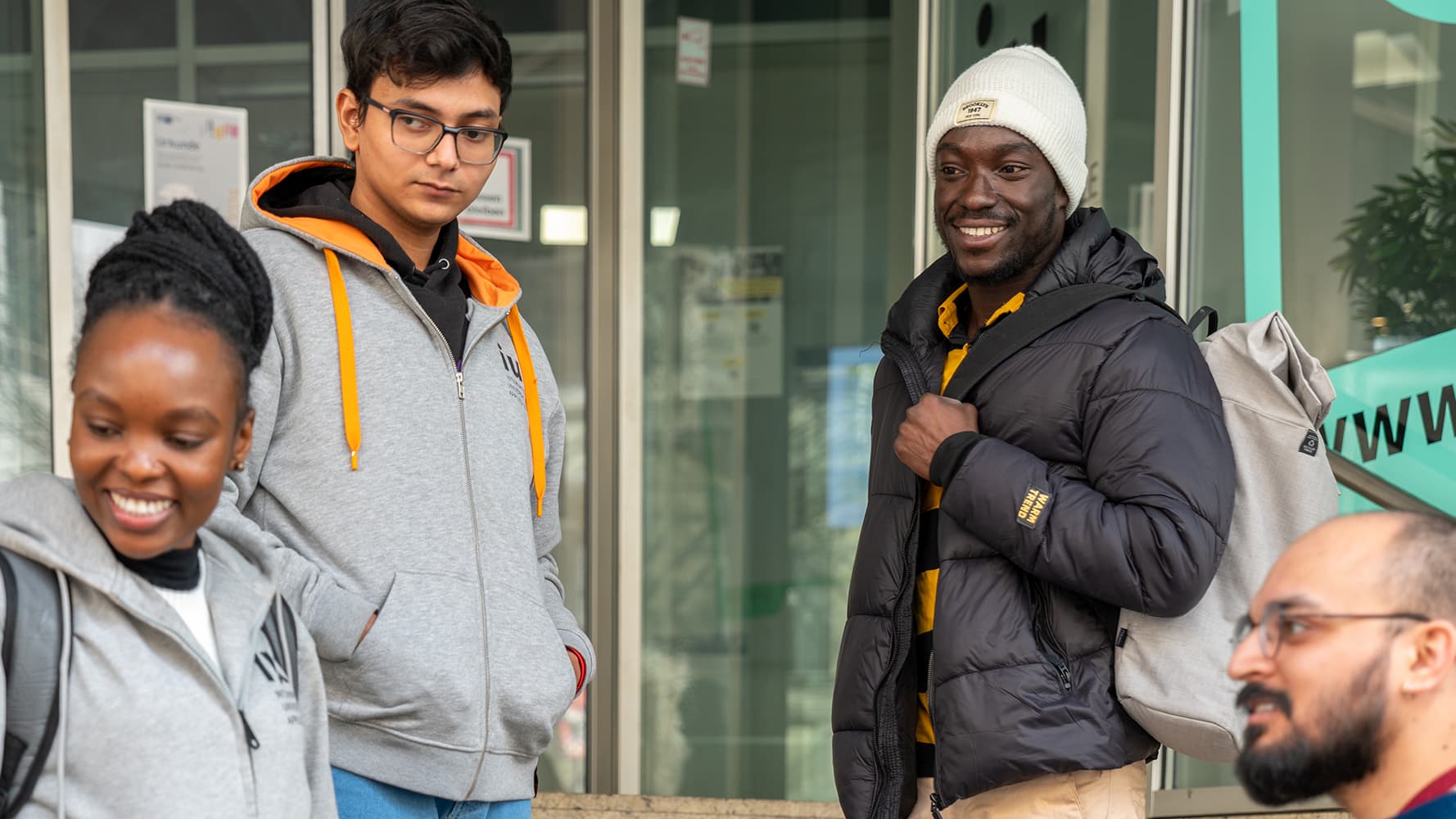 A diverse group of four students is gathered outside modern IU university building. They carry backpacks and engage in conversation, smiling and interacting.