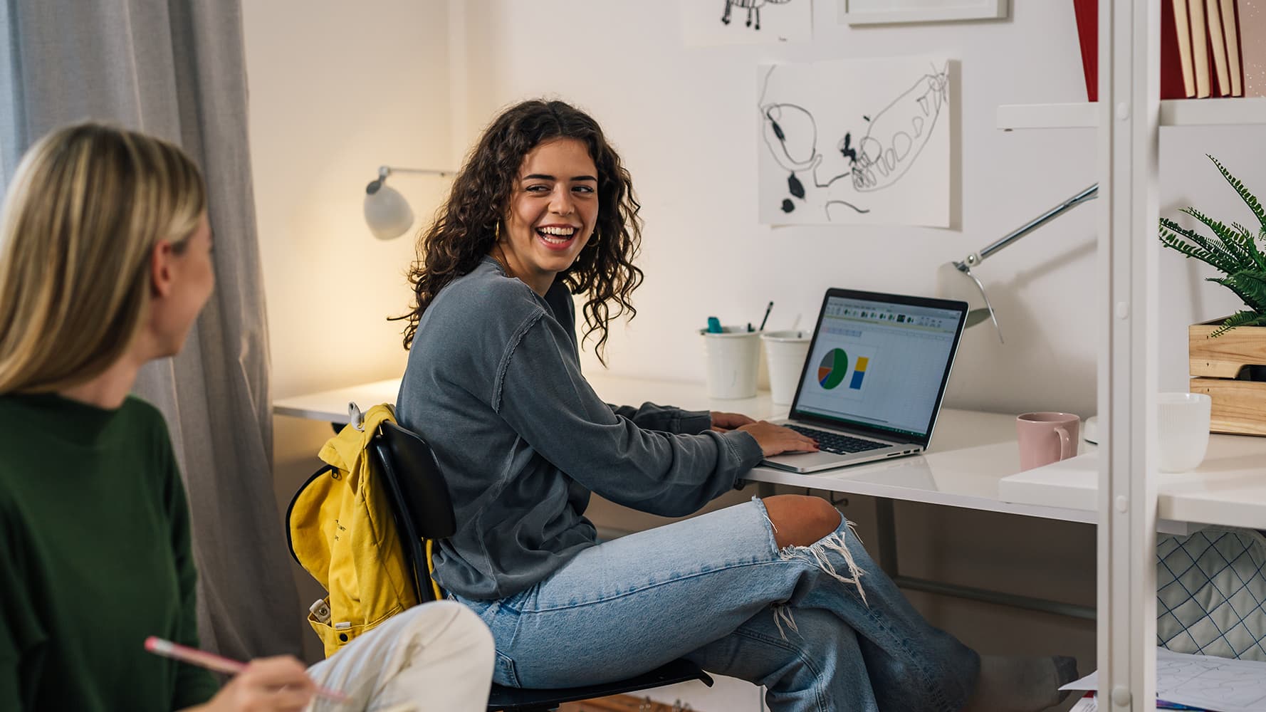 A young woman with curly hair, wearing a gray sweater, sits at a white desk working on a laptop. She is smiling and interacting with another student.