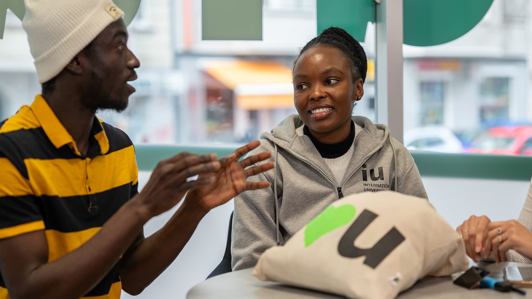 Two students sit in a university café, engaged in a lively conversation. One student wears a yellow and black striped shirt, while the other, wearing an IU International University hoodie.