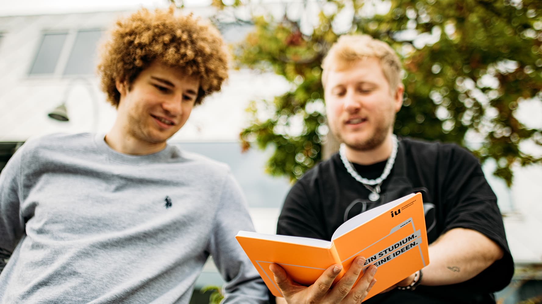 Two young students are outside looking at an open, orange, IU-branded notebook.