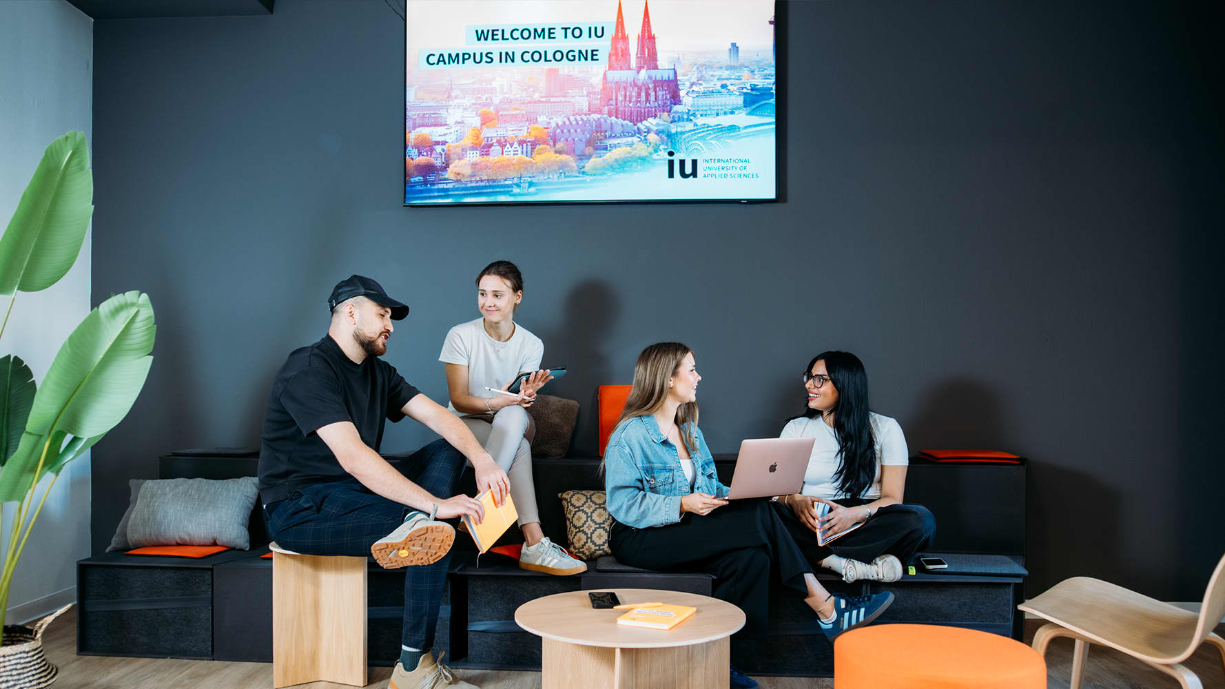 Four students are sitting casually in a modern seating area, chatting and working together. In the background, a screen displays the message 'Welcome to the IU Campus in Cologne'.
