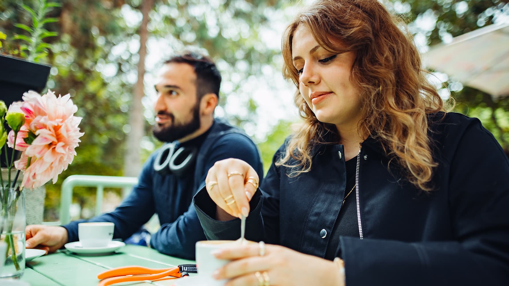 Two students sitting at an outdoor café table with flowers and coffee cups.