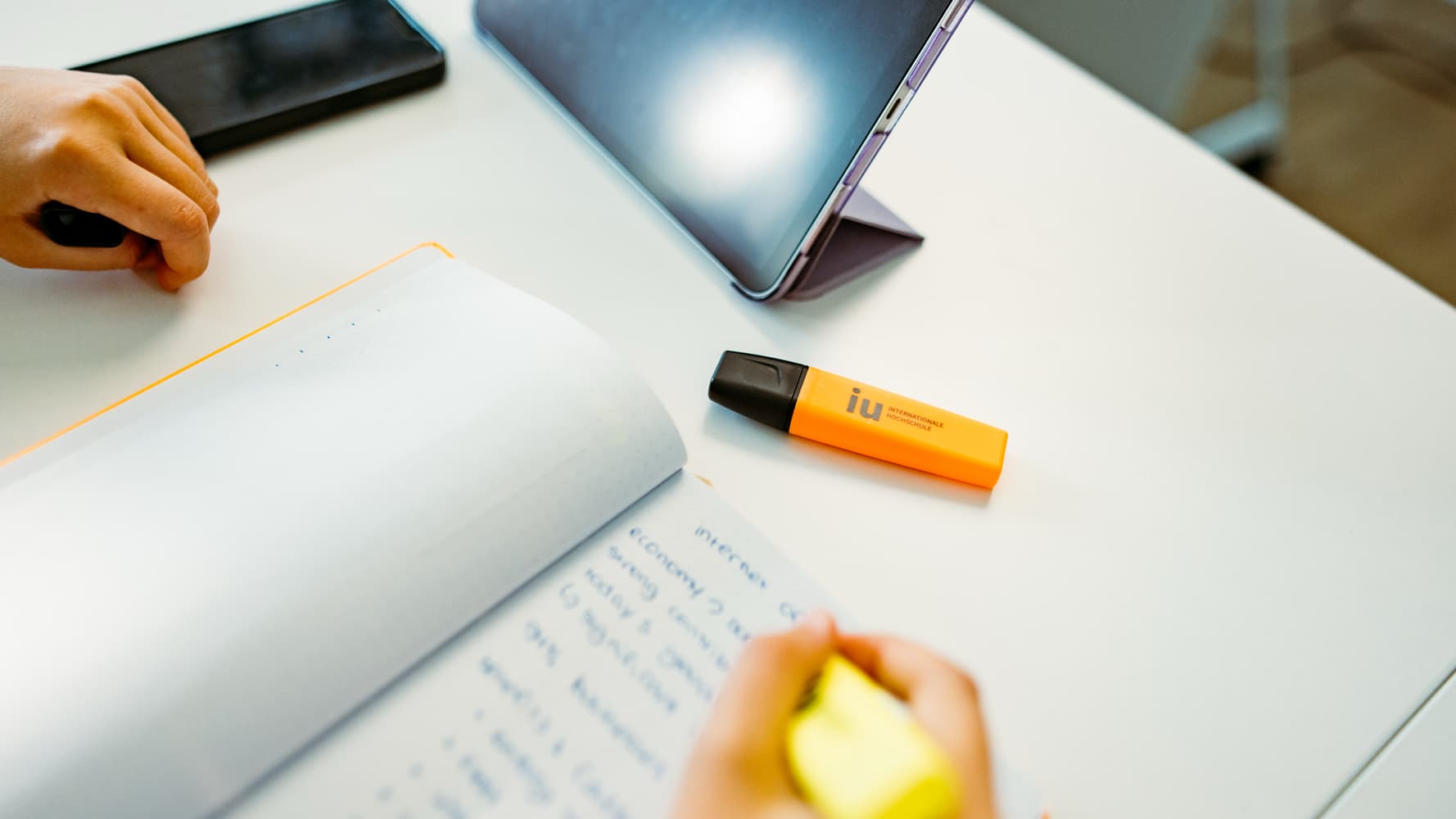 A student making study notes in a notebook with an orange branded IU International University of Applied Sciences highlighter.