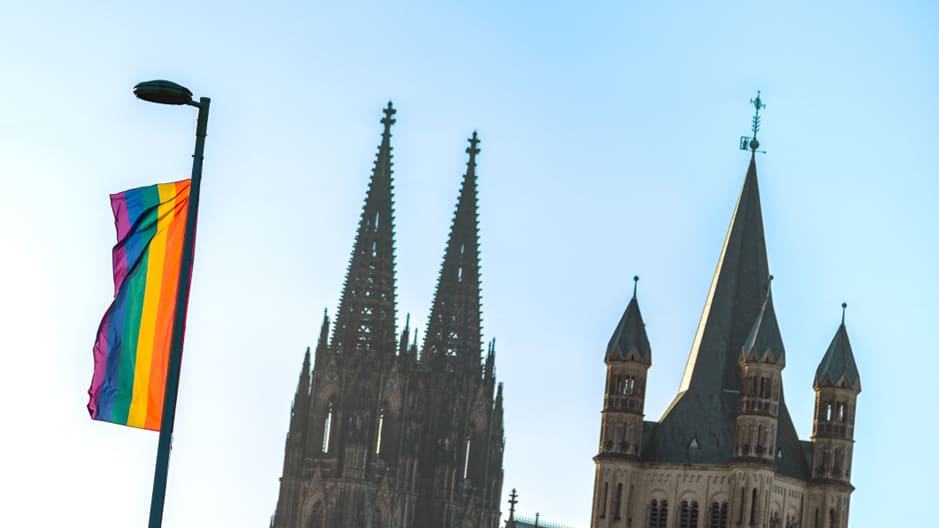 A rainbow flag waves from a lamppost against a clear blue sky, with the Cologne cathedral in the back.