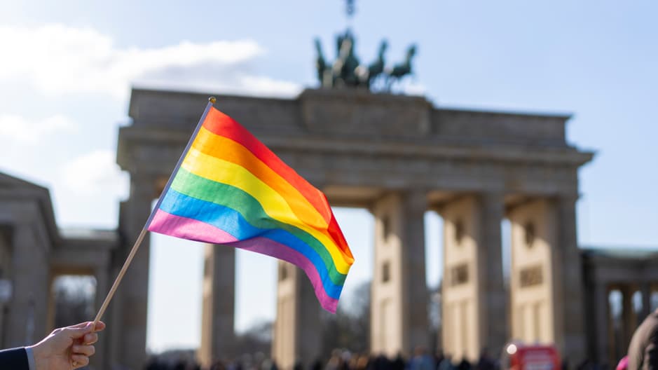 A hand holds a rainbow pride flag in front of the Brandenburg Gate in Berlin.