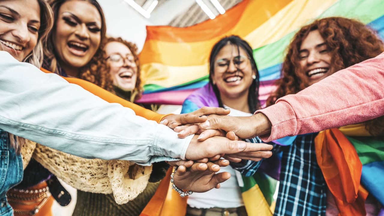 A group of friends join hands in the middle, and a LGBTQI+ community flag can be seen in the back.