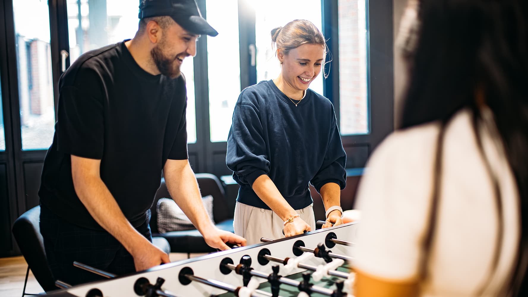 Three students are smiling and playing table football in a brightly lit room at the IU Cologne campus.