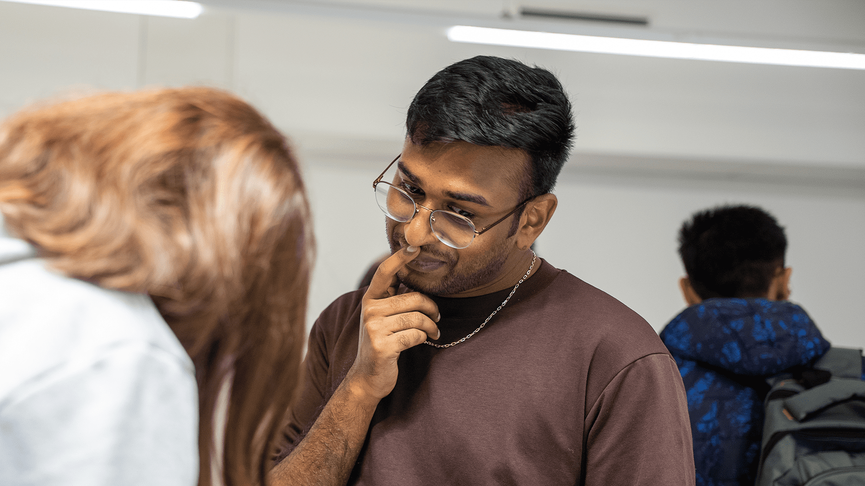 A man wearing glasses and a brown shirt is touching his chin while looking down.