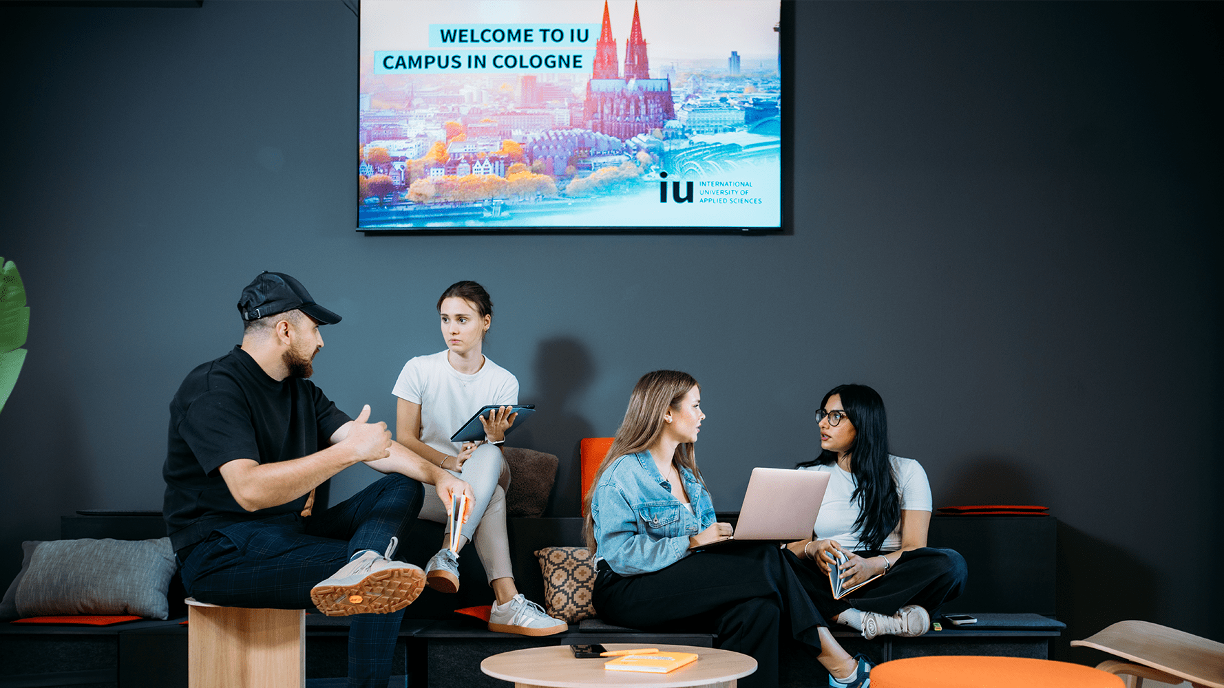 Four students sit on dark grey tiered seating in front of a large screen displaying the Cologne skyline and the text 'WELCOME TO THE IU CAMPUS IN COLOGNE'.