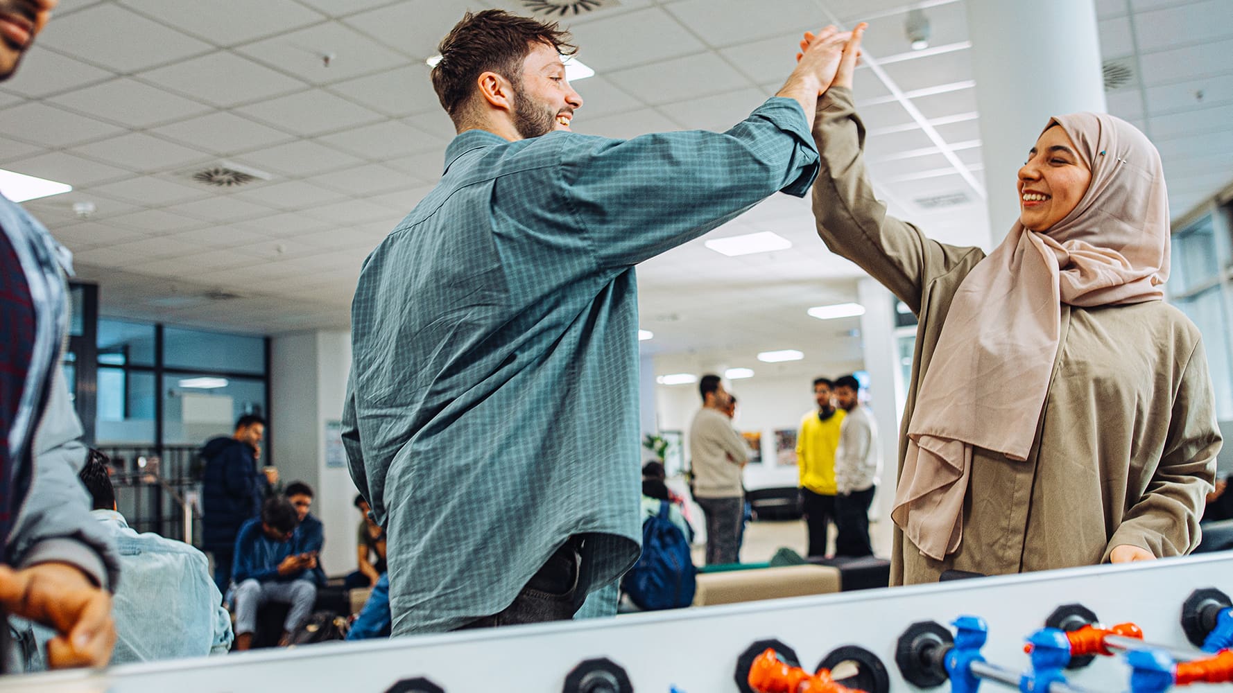 Two young students from an international university high-fiving with a smile. Other students are hanging out in the same room.