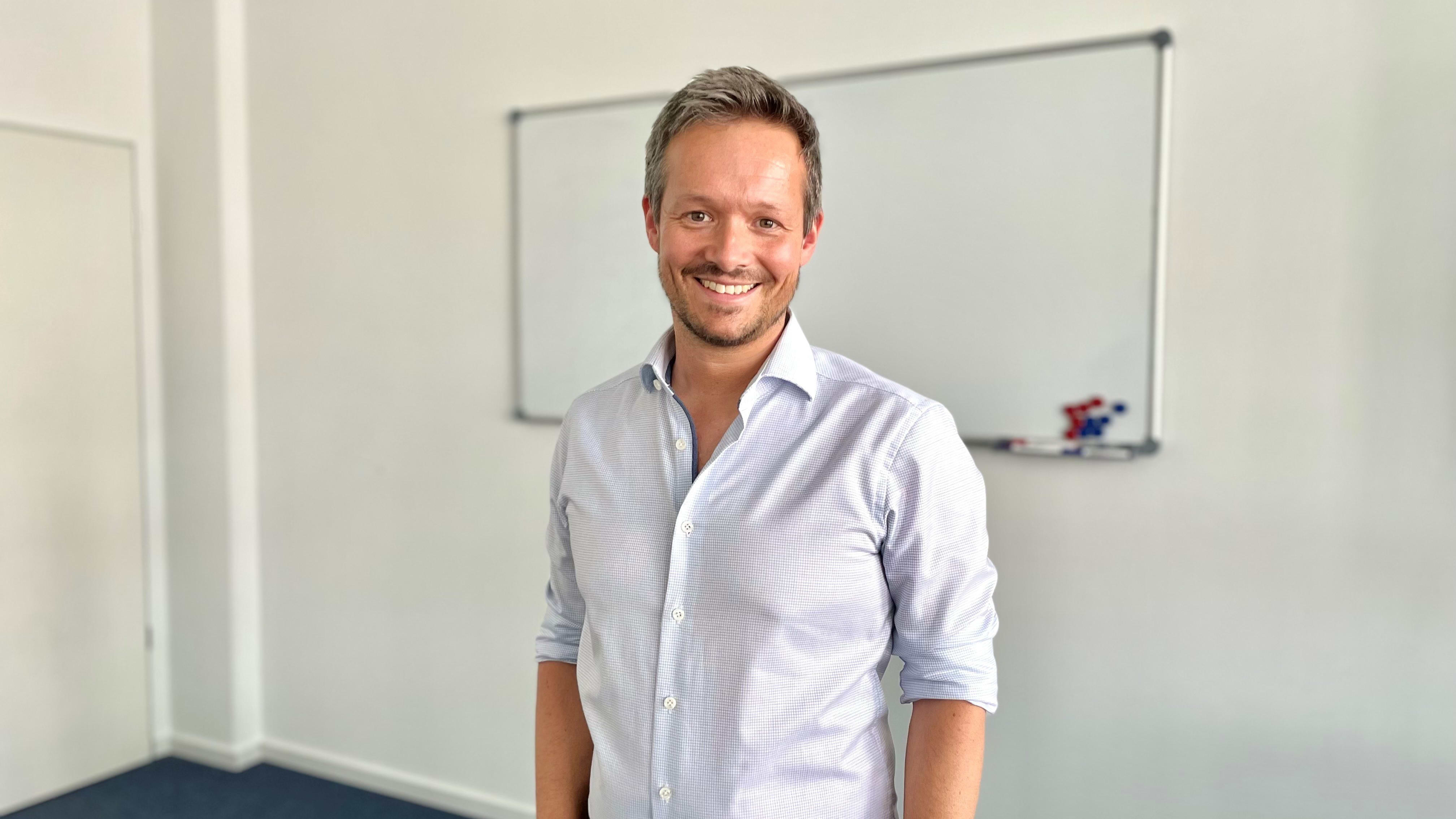 Lächelnder Mann im Business-Hemd steht in hellem Büro vor Whiteboard.
