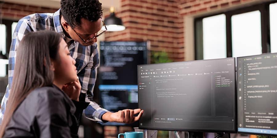Guy with glasses is pointing at the desktop and discussing tasks with his colleague as skilled ai engineers