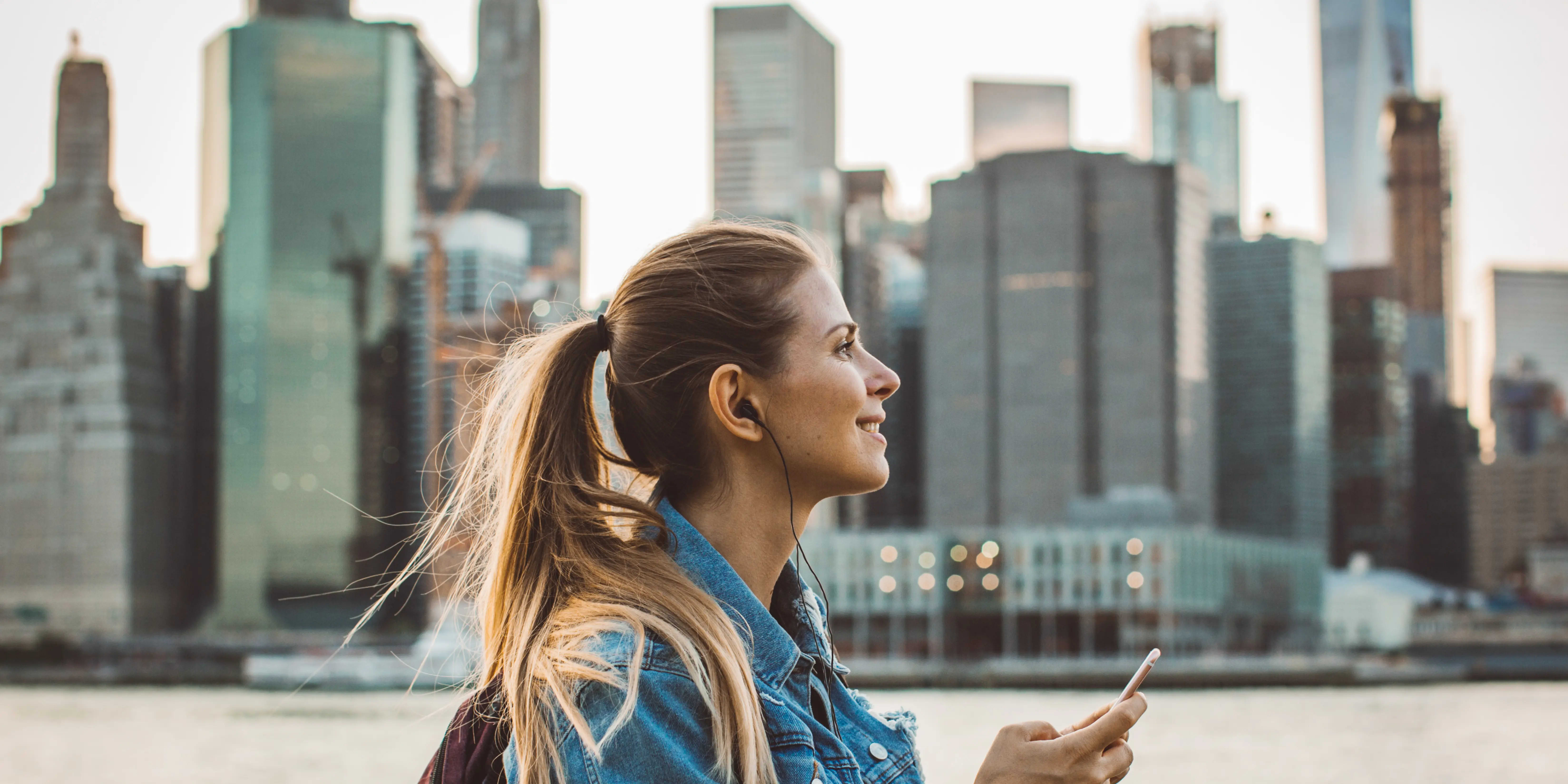 Titelbild zur Infoseite "Auslandssemester": Junge Frau mit Kopfhörern vor Skyline einer Großstadt im Ausland