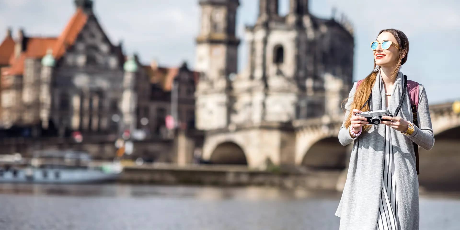 Titelbild zum Artikel "Günstige Wochenendtrips": Studentin mit Kamera vor historischer Brücke