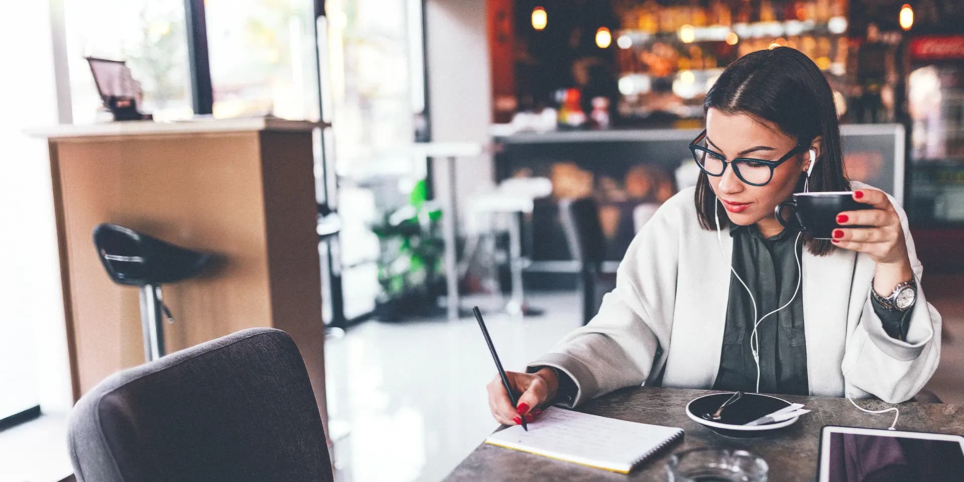 Titelbild zum Artikel "Draußen Lernen: 5 Lieblingsorte": Studentin lernt draußen im Café mit Notizen, Tablet und Kaffee.