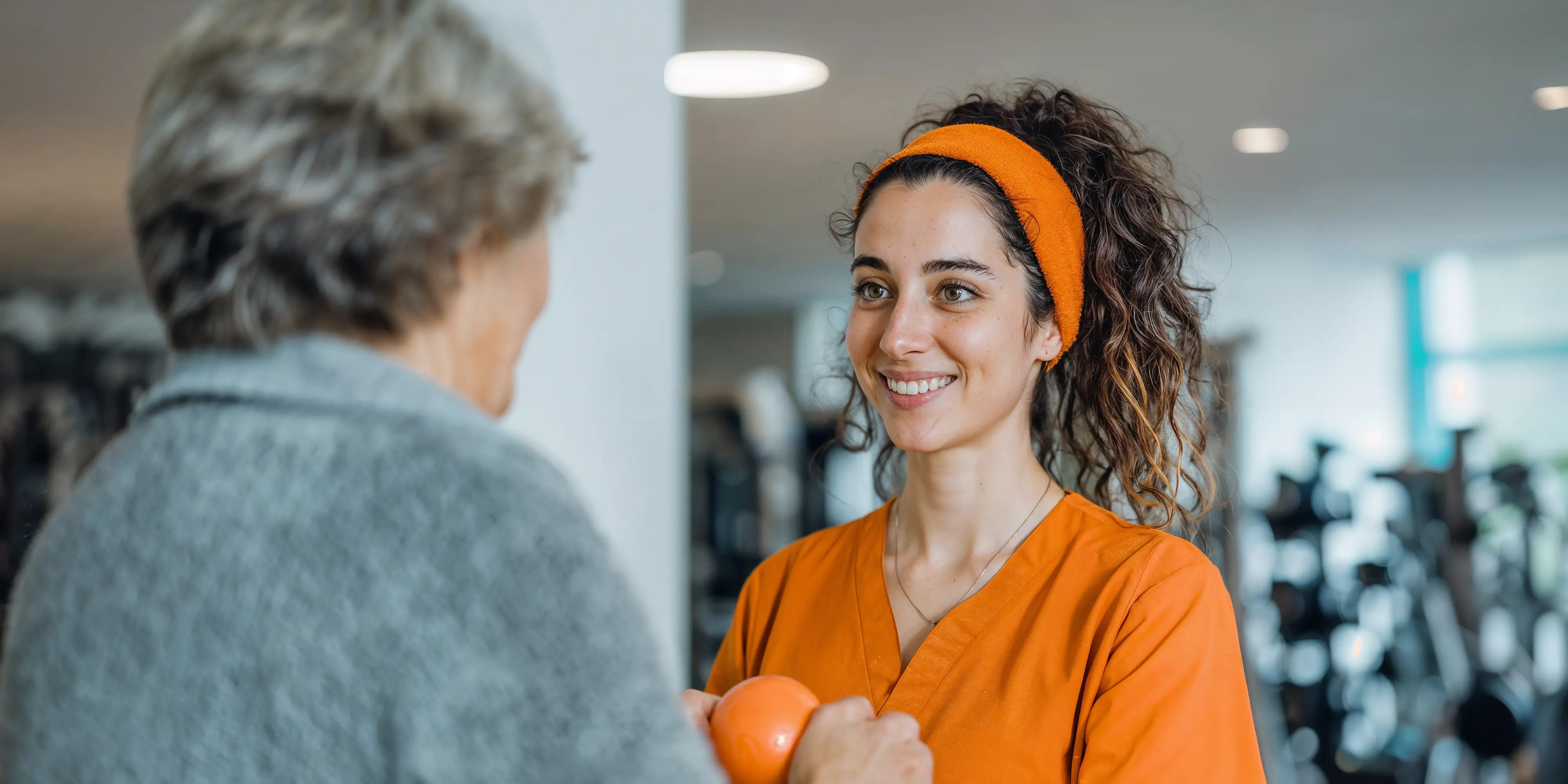 Bachelor Ergotherapie: Frau hält lächelnd einen Ball und übt mit einer älteren Person im Fitnessraum, AI-generated