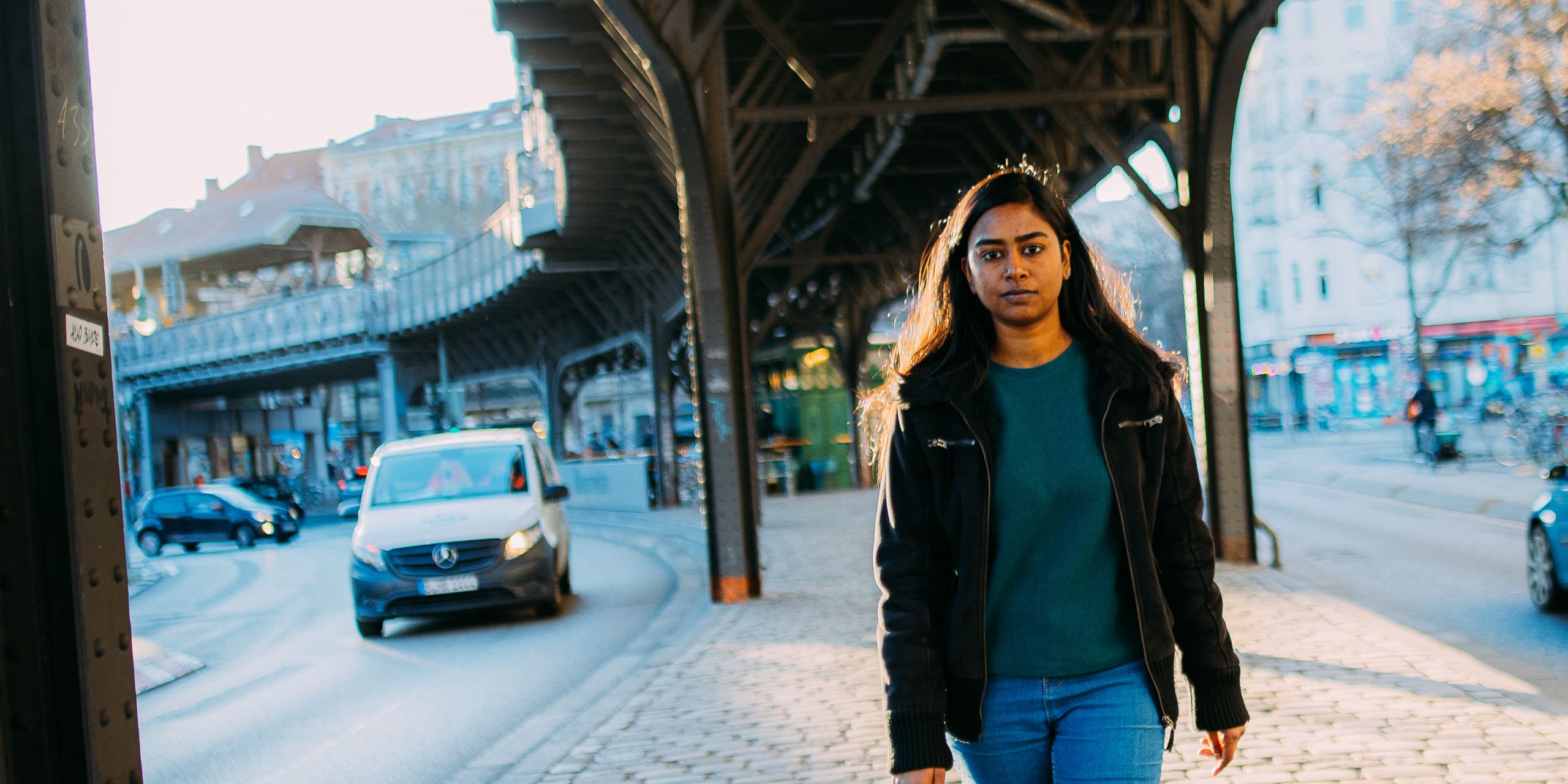 Woman in jeans and a black jacket walks under a bridge in Berlin, with cars passing nearby.