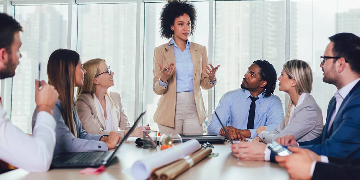 A diverse group of professionals listens attentively as a woman leads a meeting in a bright conference room, representing leadership and collaboration skills gained in a Master of Business Administration program in Germany, taught in English.