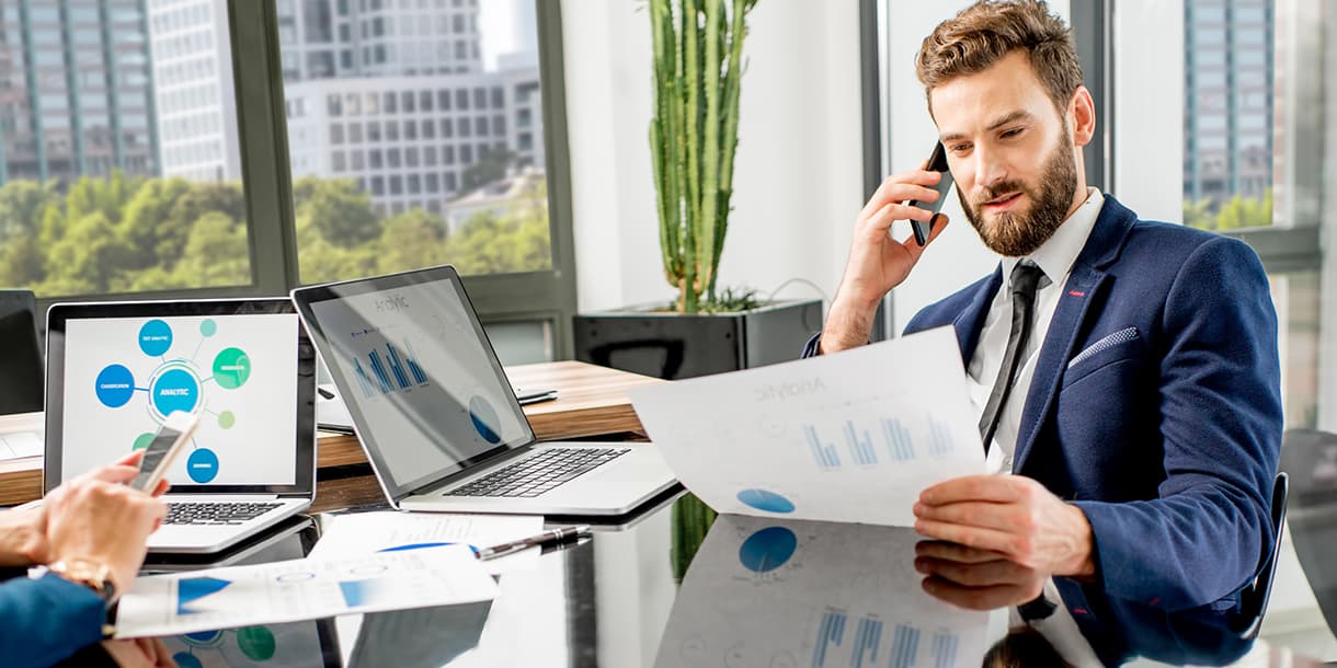 A business professional in a suit analyzes financial documents and graphs on laptops in a modern office, illustrating financial management.