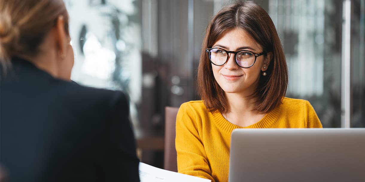 A smiling woman in glasses sits at a desk with a laptop, engaged in a discussion with another person, representing opportunities and support for international students.