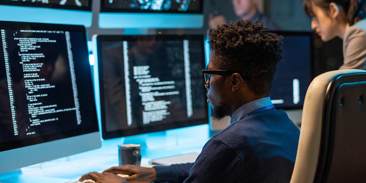 A man working intently on code displayed on a large monitor in a modern office, representing advanced technical learning in an artificial intelligence master's program in Germany.