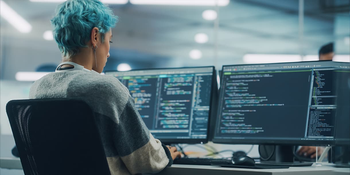 A person with blue hair works on multiple monitors displaying code in a modern office, illustrating the role of a cloud engineering consultant focused on cloud infrastructure and software development.