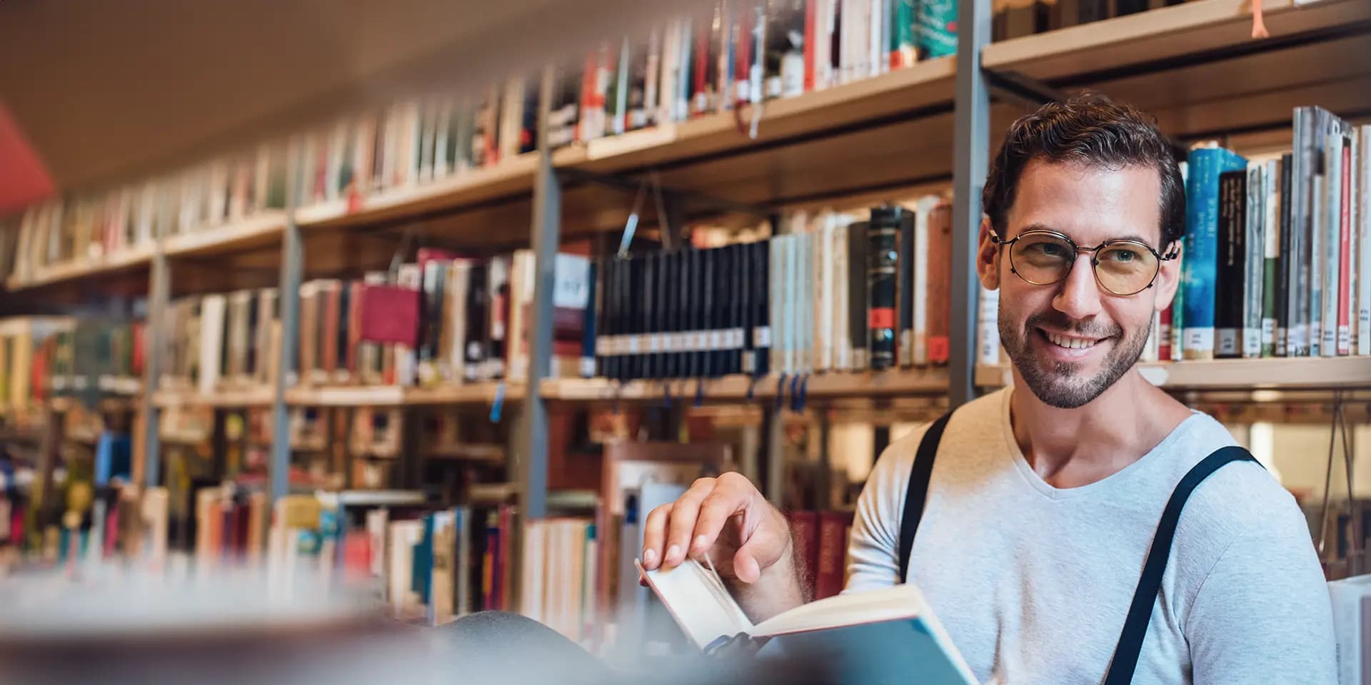 Titelbild für Artikel "Welche Doktortitel gibt es?": Mann mit Brille und Buch in Bibliothek