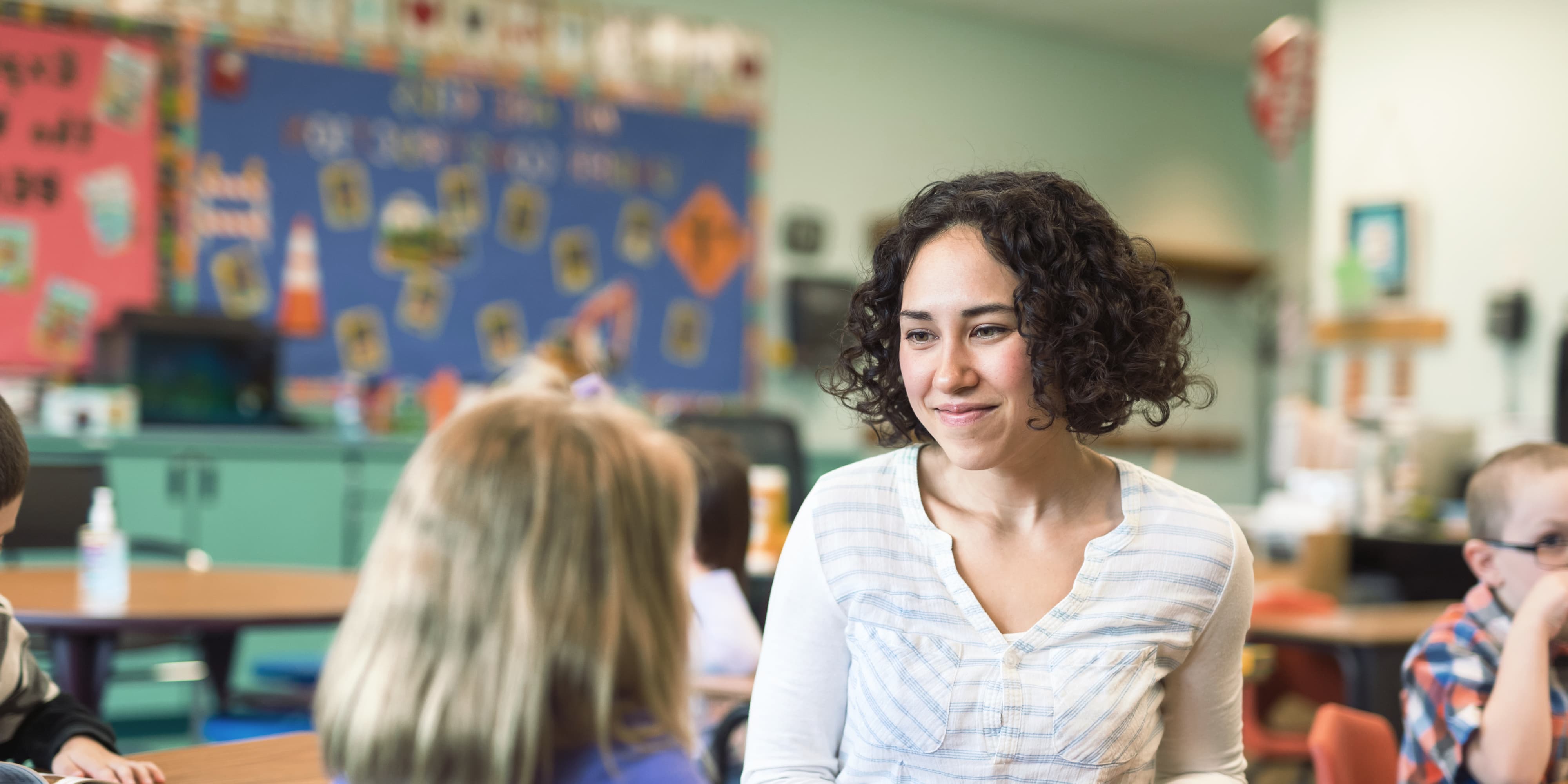 Titelbild für Infoseite "duales Studium Kindheitspädagogik": Pädagogin sitzt mit Kind am Tisch in einer Schule, AI-generated
