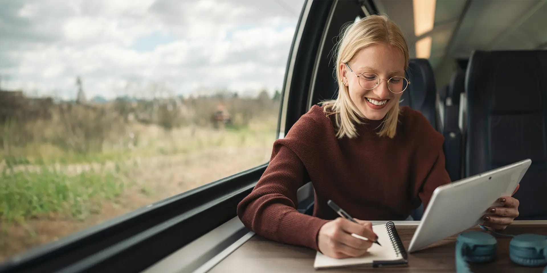 Titelbild für Artikel zum Teilzeitstudium: Lächelnde Studentin sitzt mit Tablet und Notizbuch in der Bahn
