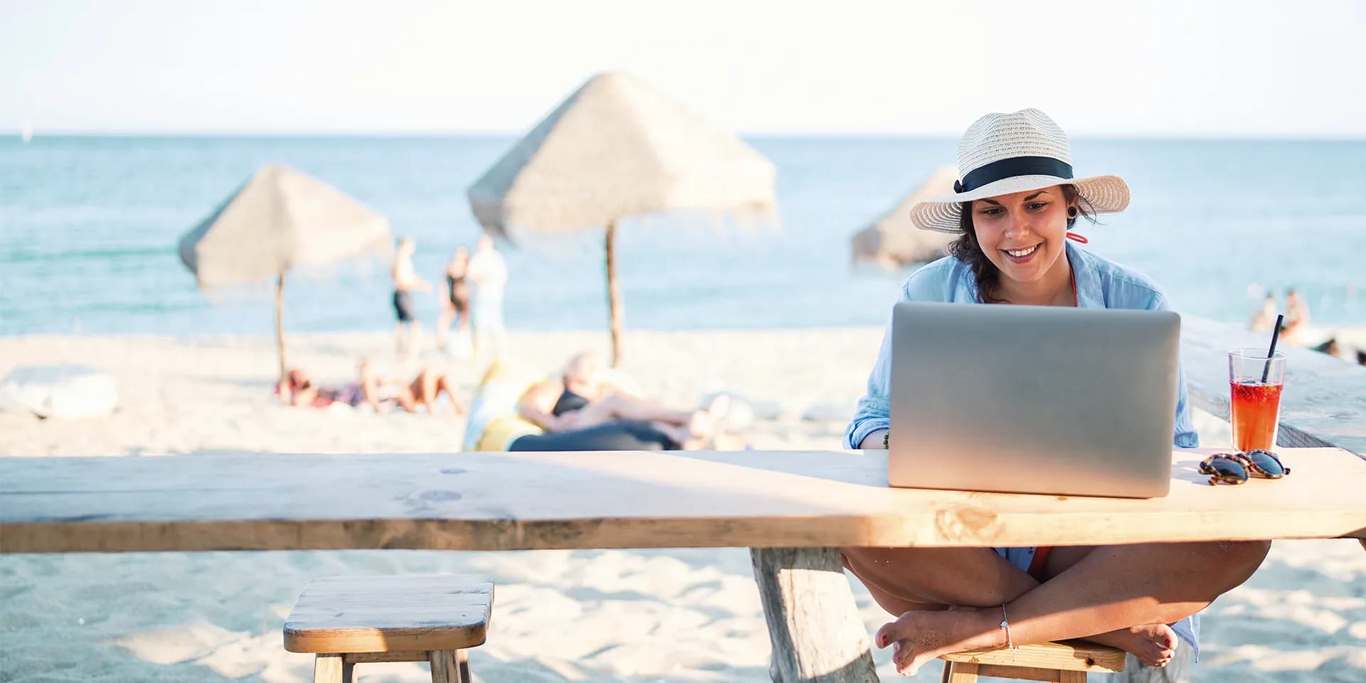 Titelbild für Artikel "Workation": Frau sitzt am Strand vor dem Meer und arbeitet am Laptop