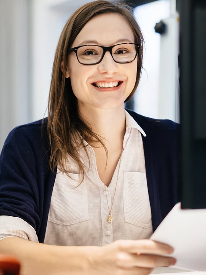 A smiling professional woman in an office setting holds documents and works at a computer, representing steps to secure a job in Germany.