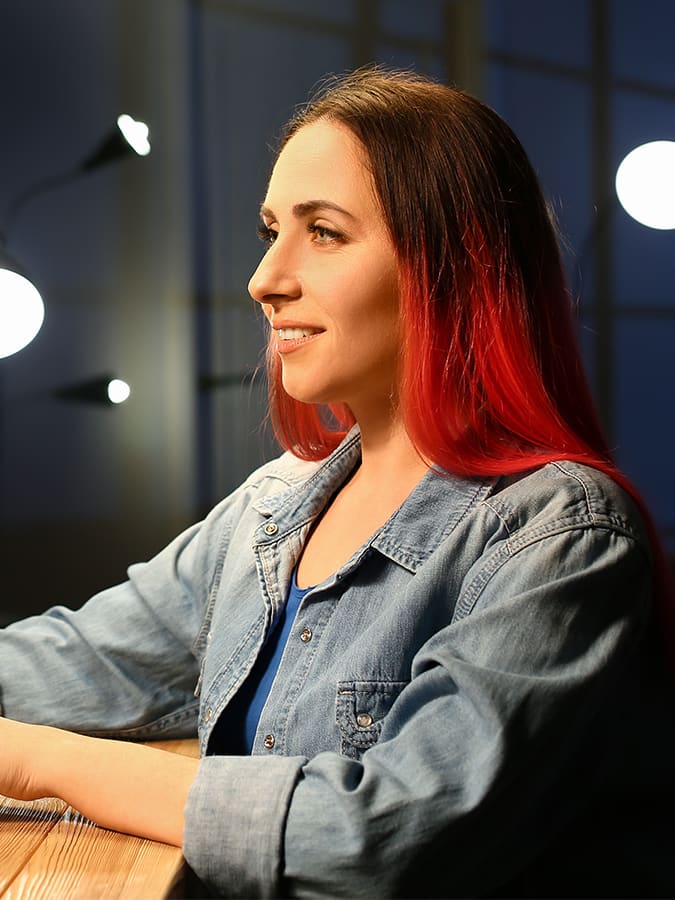 A young woman with red hair works at a computer displaying code in a dimly lit office, representing the steps to become an AI engineer through hands-on programming.