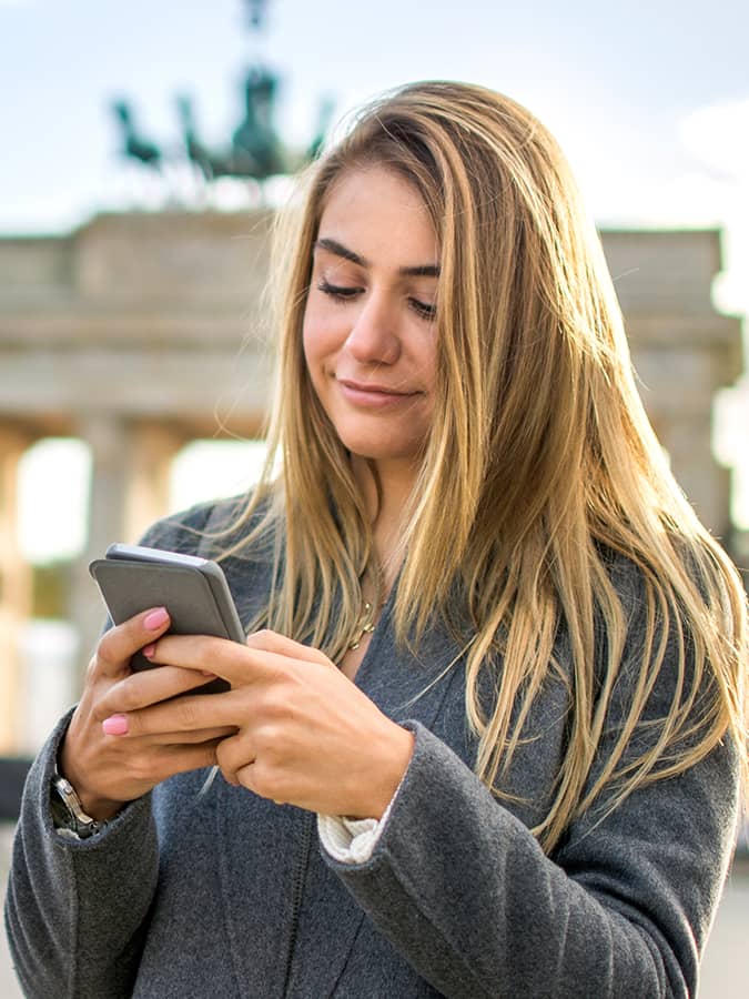 A young woman uses her smartphone in front of the Brandenburg Gate in Berlin, representing the cultural and educational advantages of studying in Germany.