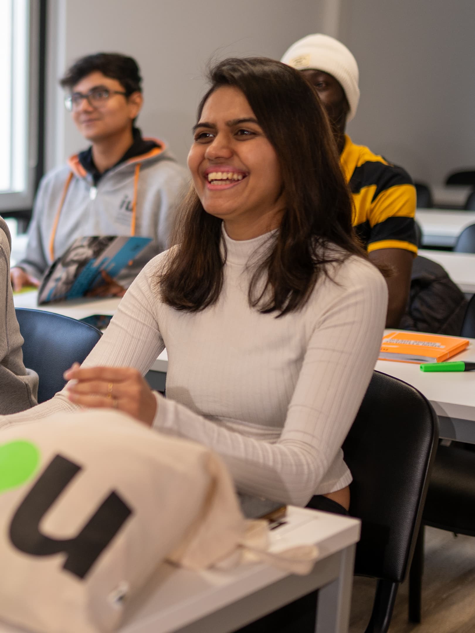 A diverse group of students is engaged in a discussion, with a young woman in a white sweater writing on a flipchart.