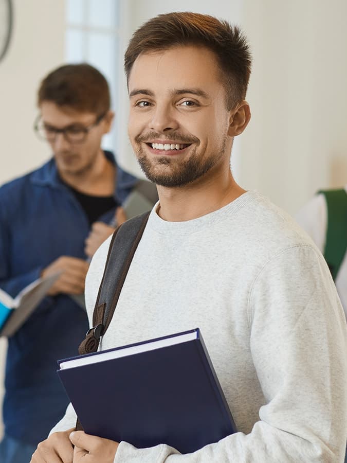 A smiling student holding a notebook stands in a study space with other students in the background, representing the community and learning environment at private universities in Germany.